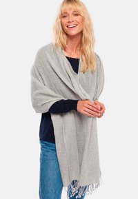 Femme aux cheveux blonds souriante, portant un pull marine, un jean bleu et un châle gris clair à franges drapé sur ses épaules.