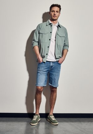 Chemise boutonnée vert clair sur un t-shirt blanc, assortie à des shorts en denim avec les manches retroussées et des baskets vertes sur un modèle devant un mur neutre.