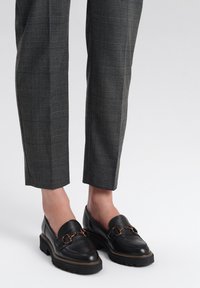 Czarne skórzane mokasyny z złotym łańcuchowym akcentem, z teksturowanym wykończeniem i grubą, żłobioną podeszwą, w zestawieniu z szarymi spodniami w kratę.