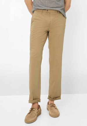 Mann trägt beige Chinos mit hochgekrempelten Saumen, kombiniert mit hellbraunen Wildleder-Schnürschuhen und einem gemusterten, eingesteckten Hemd.