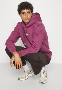 Felpa borgogna con coulisse e logo, abbinata a pantaloni in velluto a coste marroni e scarpe da ginnastica chiare. Tessuto liscio, vestibilità casual.