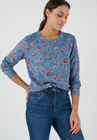 Camicia a maniche lunghe con motivo floreale blu e collo tondo, caratterizzata da fiori multicolori su un tessuto di cotone misto dalla texture interessante. Indossata con jeans di denim.