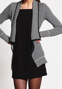 Schwarz-weiß gestreifter Cardigan mit offener Vorderseite und strukturiertem Stoff. Mit langen Ärmeln und Taschen, über einem schwarzen Kleid getragen.