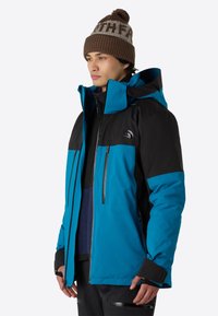 Blå och svart vinterjacka med huva, med dragkedjefickor, en texturerad inredning och justerbara ärmändar. Bärs med en brun mössa.