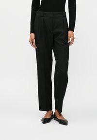 Μαύρο tailored παντελόνι με ψηλό μέσο, πιέτες και ίσια γραμμή. Το ύφασμα φαίνεται λείο με ματ φινίρισμα. Επίπεδες μαύρες γόβες.