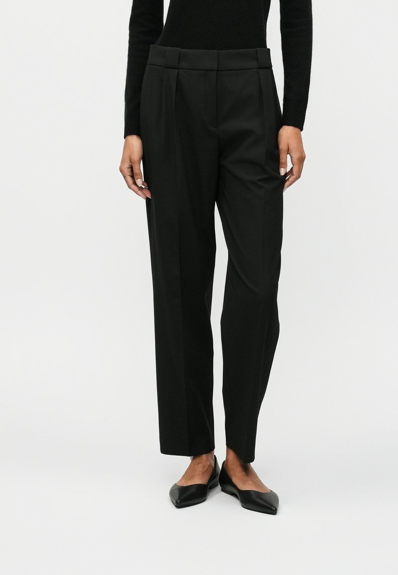 Μαύρο tailored παντελόνι με ψηλό μέσο, πιέτες και ίσια γραμμή. Το ύφασμα φαίνεται λείο με ματ φινίρισμα. Επίπεδες μαύρες γόβες.