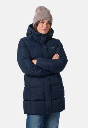 Une femme porte un manteau matelassé bleu marine à capuche et un bonnet en maille mauve. Elle se tient debout, les bras croisés, sur un fond blanc uni.