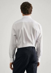 Chemise blanche à manches longues avec un col classique, texture lisse et poignets boutonnés, portée avec un pantalon sombre à motifs. Vue de dos.