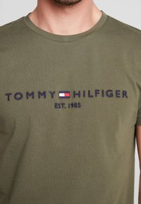 Mand iført olivengrøn T-shirt med "Tommy Hilfiger" logo og rød, hvid og blå flagemblem broderet på brystet.