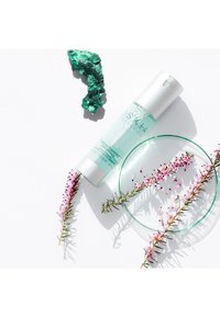 Vasetto di vetro trasparente con un coperchio argentato, contenente un liquido azzurro chiaro. Accenti floreali circostanti e una roccia minerale verde arricchiscono la scena.