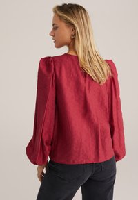Blusa cremisi in tessuto testurizzato, con scollatura tondeggiante e maniche a sbuffo. Abbinata a jeans scuri, che mostrano un fit rilassato e casual.
