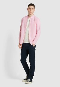 Camicia a maniche lunghe rosa chiaro con bottoni, sovrapposta a una T-shirt bianca, abbinata a pantaloni blu scuro e sneakers nere con suola bianca.