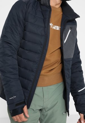 Man wearing a navy quilted jacket with zippered pocket, brown shirt, and light green pants, hands relaxed by sides.