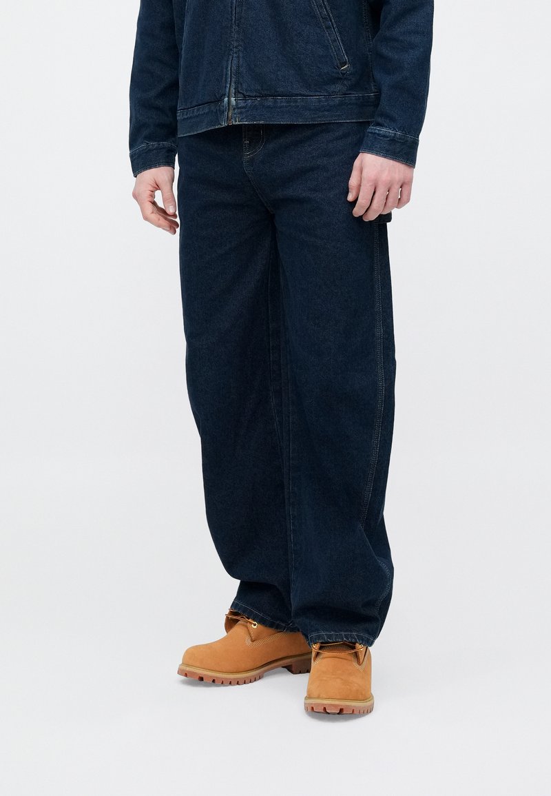 Veste en denim bleu foncé et jean ample assortis de bottes de travail en cuir beige, présentant une surface texturée et des détails de couture visibles.