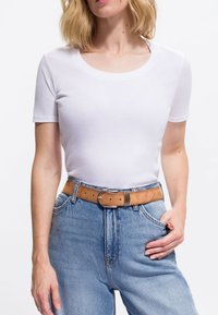 Frau in einem eng anliegenden weißen Kurzarm-T-Shirt, das in eine hoch taillierte hellblaue Jeans mit einem hellbraunen Ledergürtel gesteckt ist.