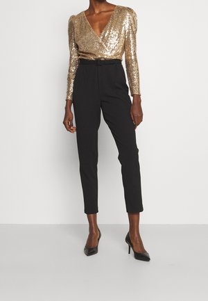 Persoon in een gouden pailletten top met lange mouwen, zwarte high-waist tapered broek en zwarte hoge hakken, staande tegen een witte muur.