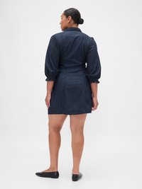 Abito in denim, blu scuro, con maniche a sbuffo e colletto. Presenta una vita stretta e una gonna svasata, mostrato di spalle, con scarpe nere piatte.