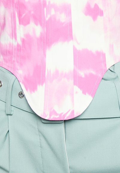 Corselete em tie-dye rosa e branco sobre calças altas de cintura azul claro, apresentando uma textura suave e botões de pressão prateados na cintura.