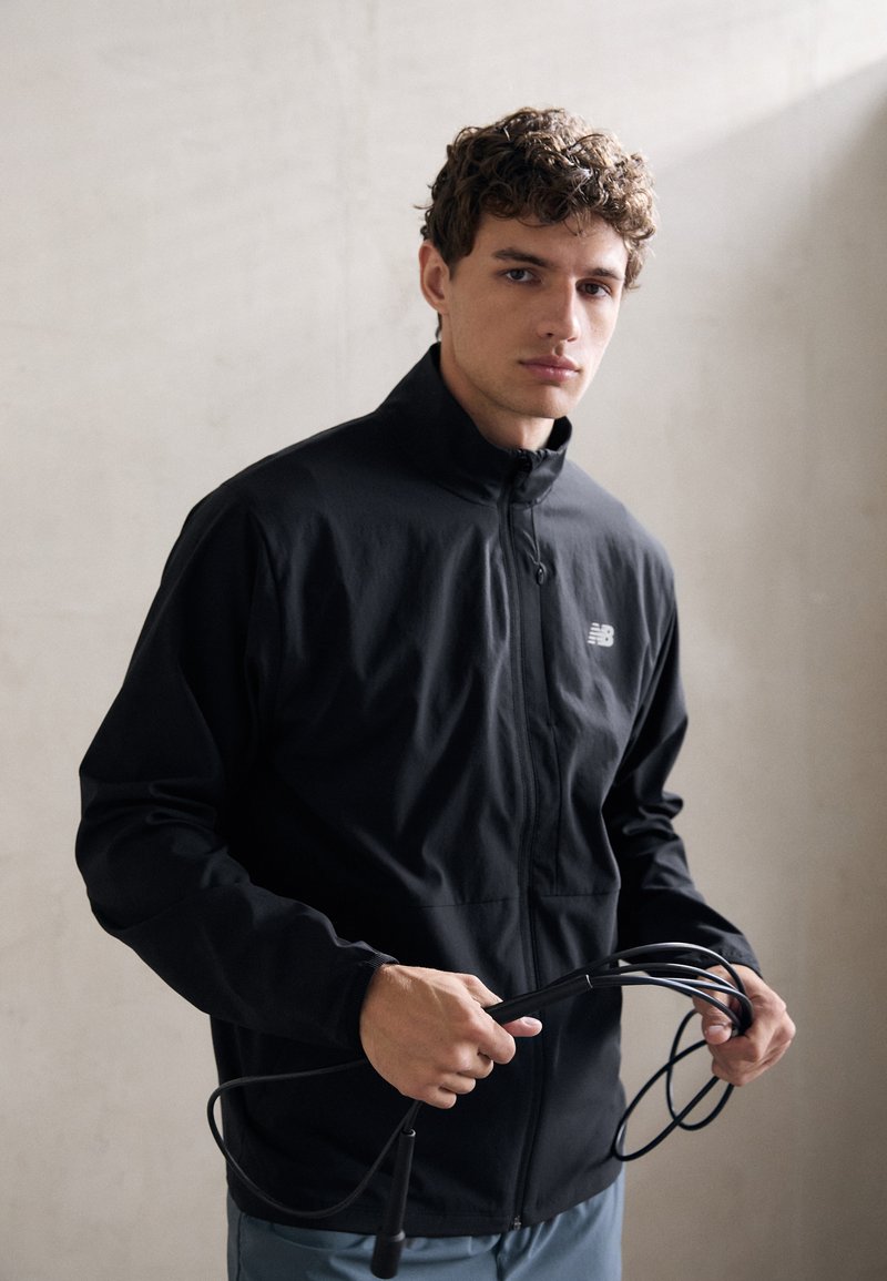 Giacca sportiva nera con zip e colletto alto, indossata da un giovane che tiene una corda per saltare. Tessuto liscio e leggero con un design aderente.