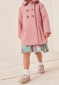 Toddler wearing pink wool coat with gold buttons, floral dress, white frilly socks, and pink shoes, walking on pale wooden floor.
