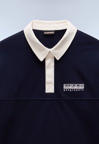 Camisa polo azul marino con un cuello crema, que presenta un detalle de punto cruz y un logotipo bordado de "Napapijri geographic" en el pecho.
