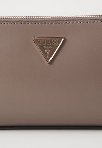 Hnědá peněženka z umělé kůže s trojúhelníkovým kovovým logem. Obsahuje zapínání na zip a vyšívané detaily pro zvýraznění designu.