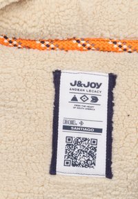Beige fleece stof met een oranje en marineblauwe geweven accent. Het aangehechte label bevat zwarte tekst, symbolen en een QR-code.