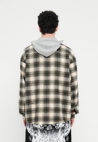 Καρό πουκάμισο φλίς σε μπεζ, μαύρο και γκρι, με γκρι κουκούλα. Το ύφασμα έχει απαλή υφή, ορατές ραφές και άνετη εφαρμογή.