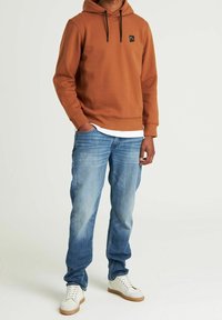 Dospělý v rezavě hnědé mikině, světlých modrých džínách a bílých teniskách, stojící s jednou rukou v kapse před jednobarevným pozadím.
