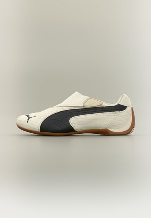 Baskets blanches sans lacets avec une bande latérale noire, une semelle marron et le logo Puma à l'avant, présentées sur un fond neutre.