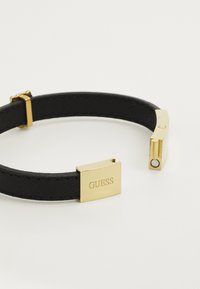Must musträni käevõru, millel on ristkülikukujuline kuldne klamber, millel on graveeritud "GUESS", esitleb sujuvat tekstuuri ja minimalistlikku disaini.