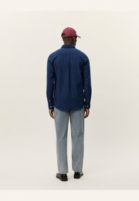 Navy blue long-sleeve shirt with button cuffs, straight cut, and back yoke detail, paired with light blue jeans and a red cap.