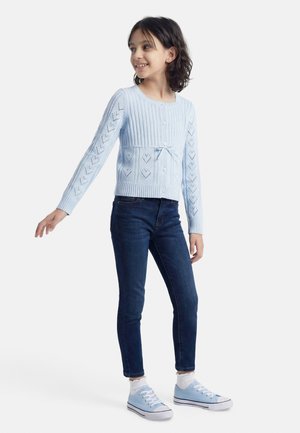 Ragazza in piedi che sorride, indossa un cardigan lavorato a maglia azzurro chiaro, jeans blu scuro, calzini bianchi e sneakers azzurro chiaro.