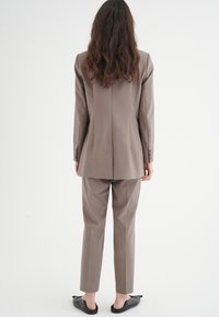 Completo beige su misura con giacca aderente e pantaloni a gamba dritta. Tessuto morbido, dettagli con spacco posteriore e scarpe nere slip-on.