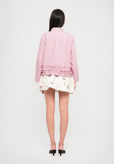 Jasnoróżowa kurtka z falbaniastym dołem, z długimi rękawami i plisą na plecach. Nosić ją na białej spódnicy z małymi kwiatowymi wzorami.