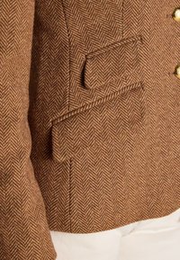 Poche de veste en tweed chevrons marron avec rabat et boutons dorés, portée sur un tissu blanc.
