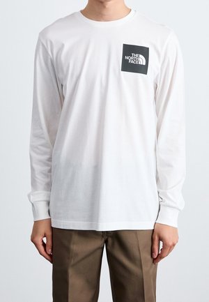 Homme portant un t-shirt blanc à manches longues avec un logo noir "The North Face" sur la poitrine et un pantalon marron.