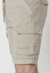 Pantaloni scurți cargo bej din bumbac, cu mai multe buzunare și accente cusute. Prezintă o margine răsucită și text embosat pe lateral.