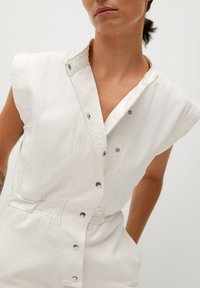 Mono de mezclilla blanca con cuello, botones a presión y mangas cortas. Presenta una cintura elástica para una silueta ajustada. Textura suave.