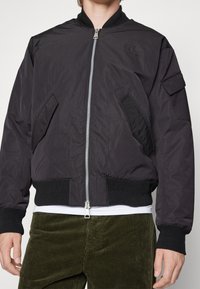 Svart bomberjacka i lättviktigt material. Har en framtill dragkedja, ribbade ärmmuddar, två sidofickor och en liten logotyp på bröstet.