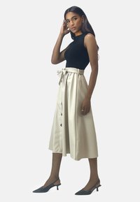 Zwart mouwloos topje gecombineerd met een crèmekleurige midi-rok met hoge taille, voorzien van een strikceintuur en knoopdetails, gedragen met zwarte slingback pumps.