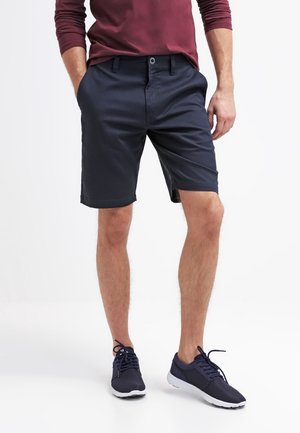 Mann trägt marineblaue Shorts, burgunderrotes Langarmshirt und marineblaue Sneakers, steht mit den Händen in den Taschen.