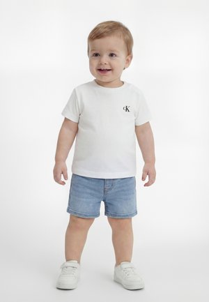 Tout-petit portant un T-shirt blanc à manches courtes avec logo, un short en jean bleu et des baskets blanches, debout et souriant sur un fond blanc.