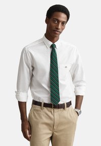 Camicia bianca con bottoni, cravatta a righe verdi e dorate, pantaloni color kaki, cintura marrone, orologio argentato, vestibilità comoda, con maniche arrotolate e dettaglio sulla tasca.