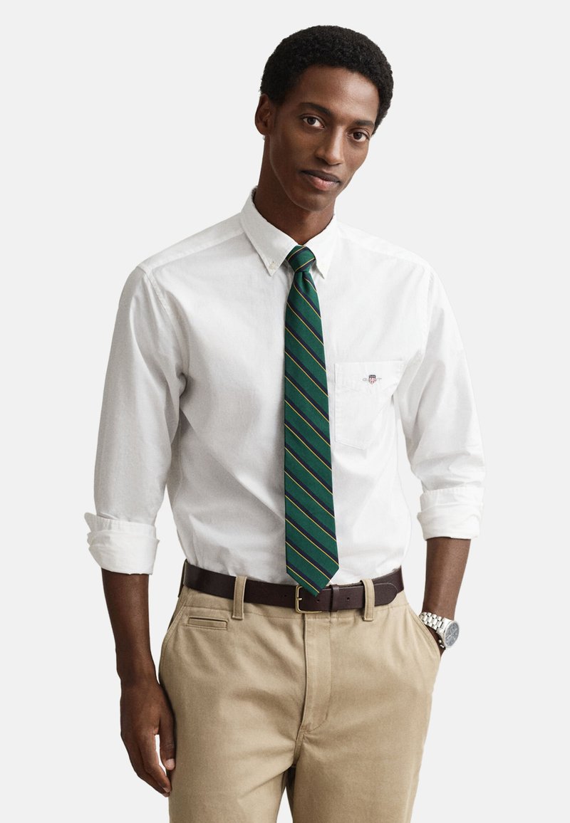 Camicia bianca con bottoni, cravatta a righe verdi e dorate, pantaloni color kaki, cintura marrone, orologio argentato, vestibilità comoda, con maniche arrotolate e dettaglio sulla tasca.