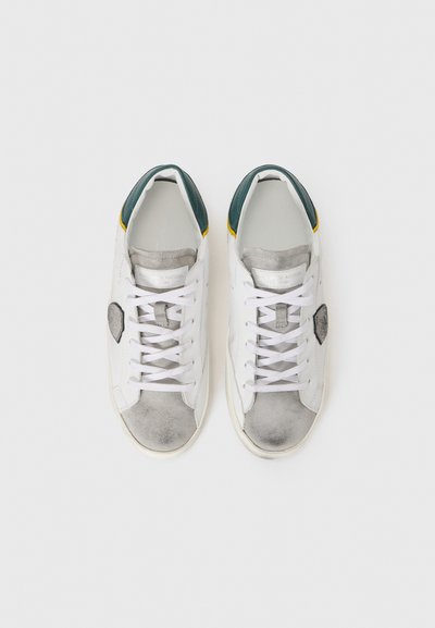 White sneakers with gray suede accents, green and yellow heel tabs, and flat white laces. Features a logo patch on the side.