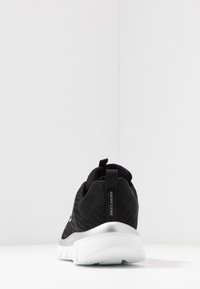 Chaussure de sport noire avec une tige en maille texturée, semelle blanche rembourrée et un accent argenté. Dispose d'une languette au talon pour un enfilage facile.