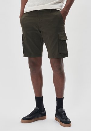 Pantaloni cargo verde scuro con tasche laterali, passanti per cintura e taglio dritto. Abbinati a sneakers nere e calze. Tessuto morbido.
