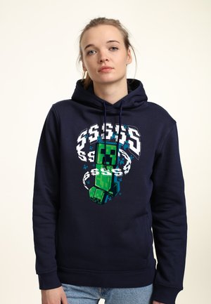 Granatowy hoodie z bawełny, z pikselowym zielonym wzorem postaci w białych akcentach oraz kapturem ze sznurkiem. Model nosi go z dżinsami.
