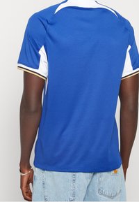 Camiseta deportiva azul con acentos blancos, mangas cortas y puños a rayas doradas; hecha de tela texturizada, con un cuello redondo y un dobladillo recto.